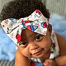 baby, bow, child, close_up, clothing, colorful, curly_hair, cute, expression, face, headband, indoors, infant, person, playful, portrait, skin, smile, soft_background, young