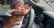 Cierra joined the competition — help win amazing prizes! baby, child, infant, car, steering_wheel, hat, knit_hat, purple_hat, hands, clothing, window, dashboard, person, vehicle, parking_lot, trees, overcast, cute, indoor, portrait