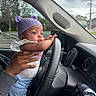 baby, child, infant, car, steering_wheel, hat, knit_hat, purple_hat, hands, clothing, window, dashboard, person, vehicle, parking_lot, trees, overcast, cute, indoor, portrait