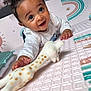 baby, child, toy, giraffe, play_mat, smiling, happy, indoor, toddler, curly_hair, white_clothing, hand, face, large_eyes, crawling, colorful, patterned, soft_toy, cute, playtime