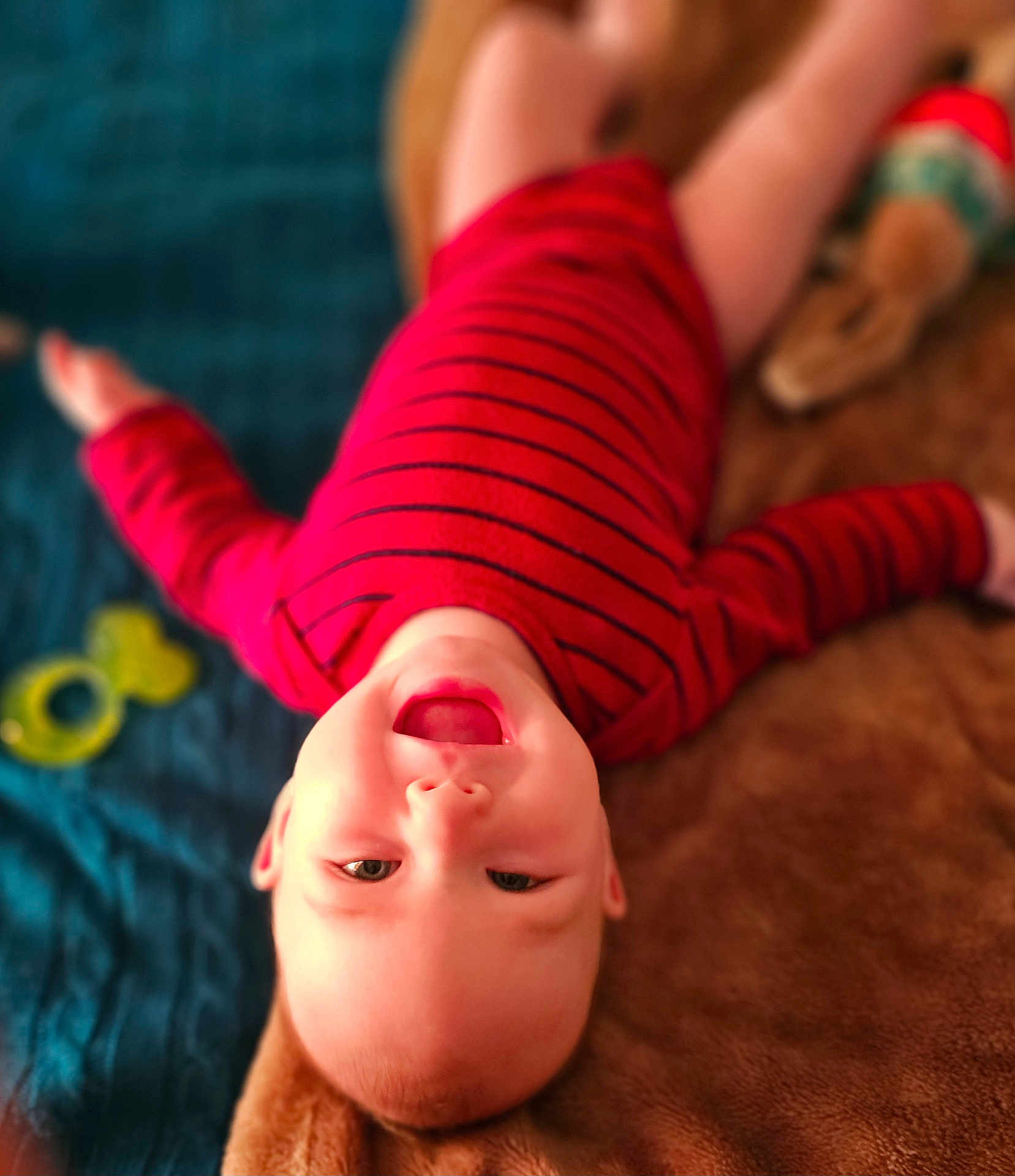 Levi is registered to the contest to win money with this photo: baby, child, smiling, red_clothing, striped_shirt, blanket, toy, blue_quilt, happy, face, head, lying_down, indoor, infant, person, cute, playful, warm, soft_texture, mouth_open