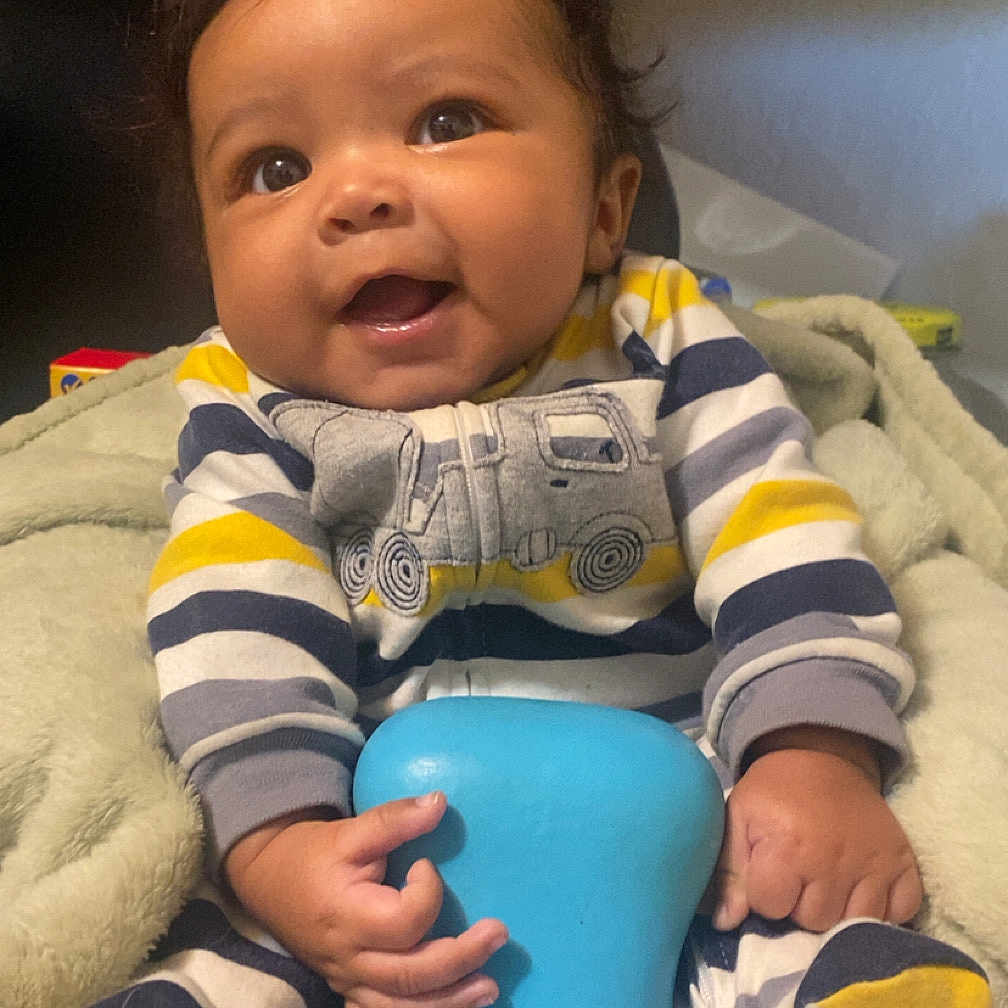 Jayceon is registered to the contest to win money with this photo: baby, child, smiling, striped_clothing, onesie, blue_toy, blanket, indoor, happy, cute, infant, young_child, sitting, face, hands, feet, furniture, warm_lighting, portrait, playful