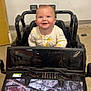 baby, child, smiling, toy_car, indoor, onesie, happy, vehicle, camouflage, floor, cardboard_box, seat, face, head, person, play, young_child, light, expression, background