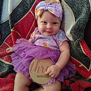 Zariah joined the competition — help win amazing prizes! baby, blanket, child, clothing, cute, face, hand, happy, headband, indoors, infant, legs, milestone, person, portrait, purple_tutu, sitting, smile, three_months, wooden_sign