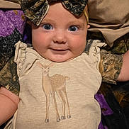 Gracelynn is registered to the contest to win money with this photo: baby, infant, smile, face, headband, bow, clothing, onesie, deer_print, blanket, purple, brown, camo_pattern, cute, lying_down, child, person, indoors, happy, portrait