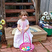 Evelyn is registered to the contest to win money with this photo: child, girl, white_dress, basket, colorful_eggs, wooden_stairs, outdoor, garden, flowers, greenery, grass, brick_path, smiling, cute, daylight, playful, spring, backyard, decorations, toddler