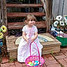 backyard, basket, brick_path, child, colorful_eggs, cute, daylight, decorations, flowers, garden, girl, grass, greenery, outdoor, playful, smiling, spring, toddler, white_dress, wooden_stairs