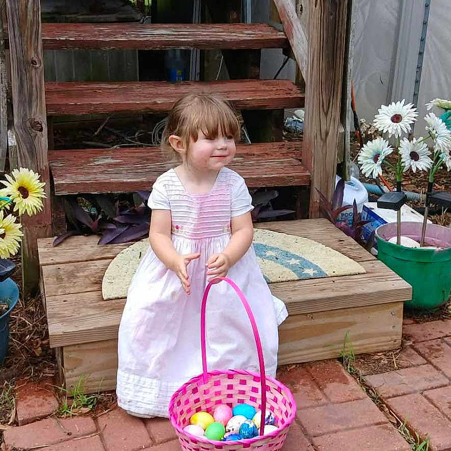 Evelyn is registered to the contest to win money with this photo: child, girl, white_dress, basket, colorful_eggs, wooden_stairs, outdoor, garden, flowers, greenery, grass, brick_path, smiling, cute, daylight, playful, spring, backyard, decorations, toddler