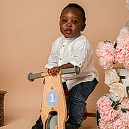 Alvin a rejoint le concours — aidez-le/la à gagner de superbes lots ! toddler, child, balance_bike, wooden_toy, white_shirt, jeans, black_shoes, flowers, pink_flowers, white_flowers, portrait, indoor, studio, smiling, cute, baby, person, toy, background, decor