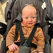 Antonn participe au concours pour gagner de l'argent avec cette photo : baby, infant, stroller, car_seat, harness, smile, corduroy, brown_outfit, bald_head, wide_eyes, rosy_cheeks, hand, buckle, seat_padding, portrait, closeup, indoor, cute, child, person