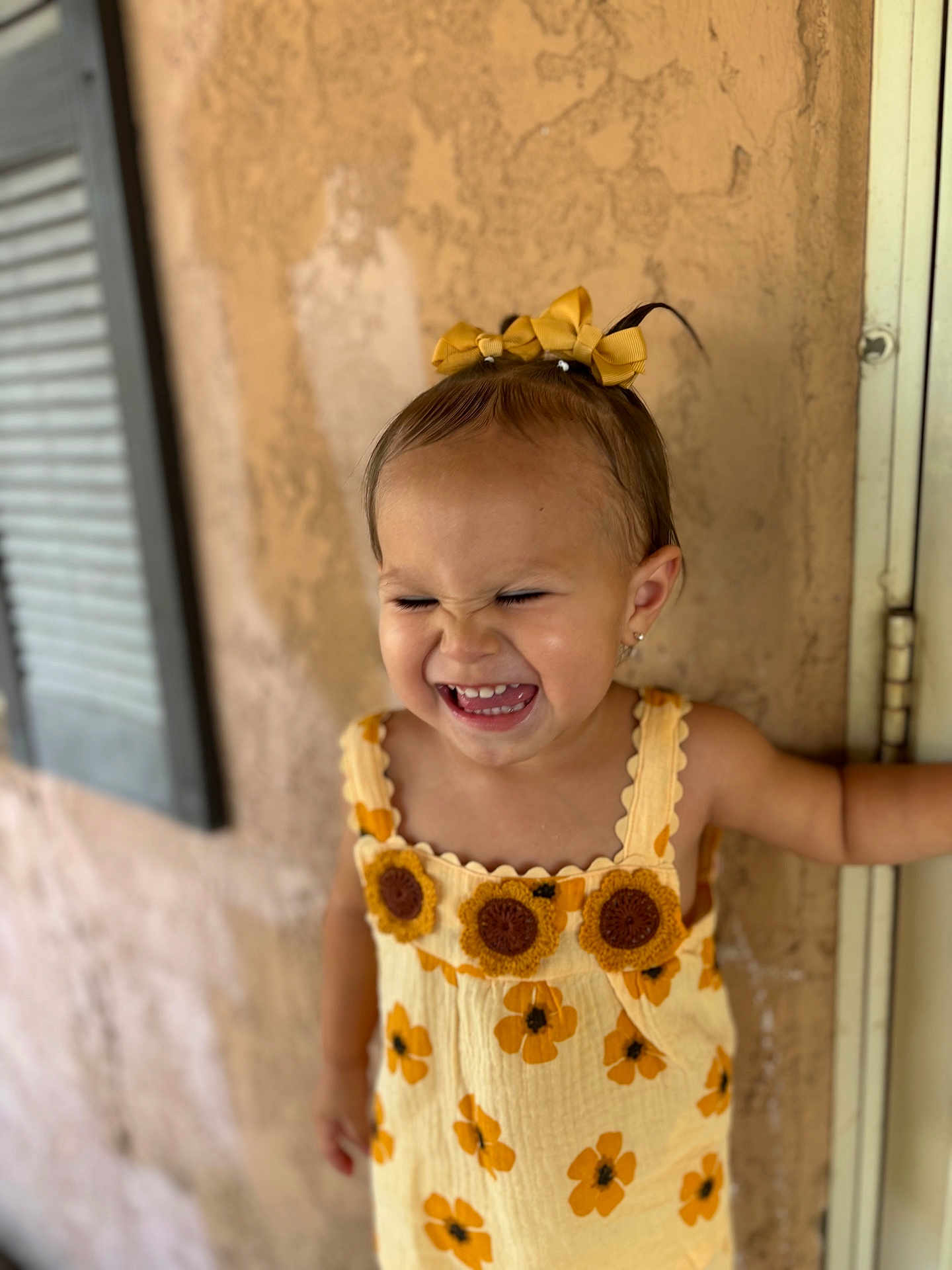 Celina is registered to the contest to win money with this photo: toddler, child, smiling, laughing, yellow_dress, sunflower_pattern, hair_bows, earrings, outdoor, wall, window_shutters, happy, cute, portrait, person, summer, fashion, texture, young_child, playful