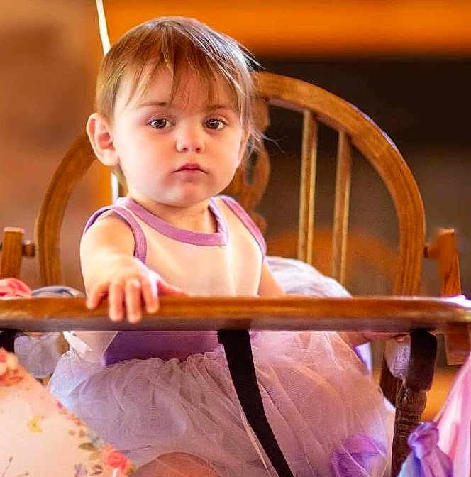 Miley joined the competition — help win amazing prizes! child, toddler, high_chair, dress, purple, white, wooden_chair, indoor, curious, face, person, young, sitting, baby, clothing, furniture, portrait, soft_light, casual, home