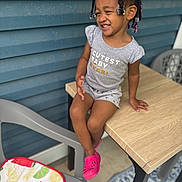 Ziva is registered to the contest to win money with this photo: toddler, child, girl, braided_hair, beads, smiling, grey_outfit, pink_shoes, wooden_table, plastic_chair, fruit_cushion, blue_wall, outdoor, sitting, happy, portrait, casual, cute, playful, daylight