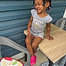 toddler, child, girl, braided_hair, beads, smiling, grey_outfit, pink_shoes, wooden_table, plastic_chair, fruit_cushion, blue_wall, outdoor, sitting, happy, portrait, casual, cute, playful, daylight