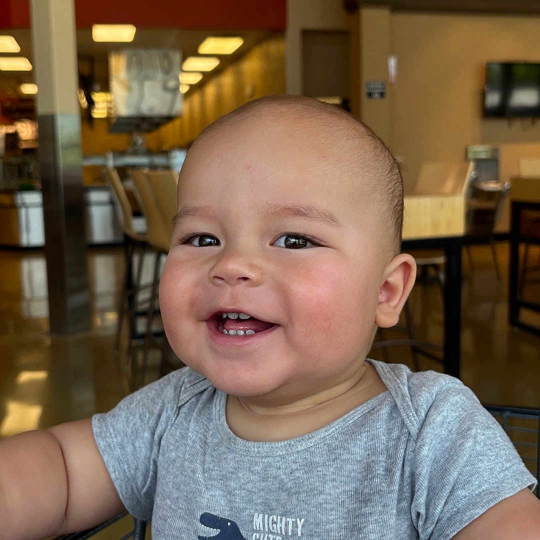 Jaden is registered to the contest to win money with this photo: arm, baby, chair, child, clothing, cute, dinosaur, face, furniture, happy, head, indoor, onesie, person, shopping_cart, skin, smile, store, table, toddler