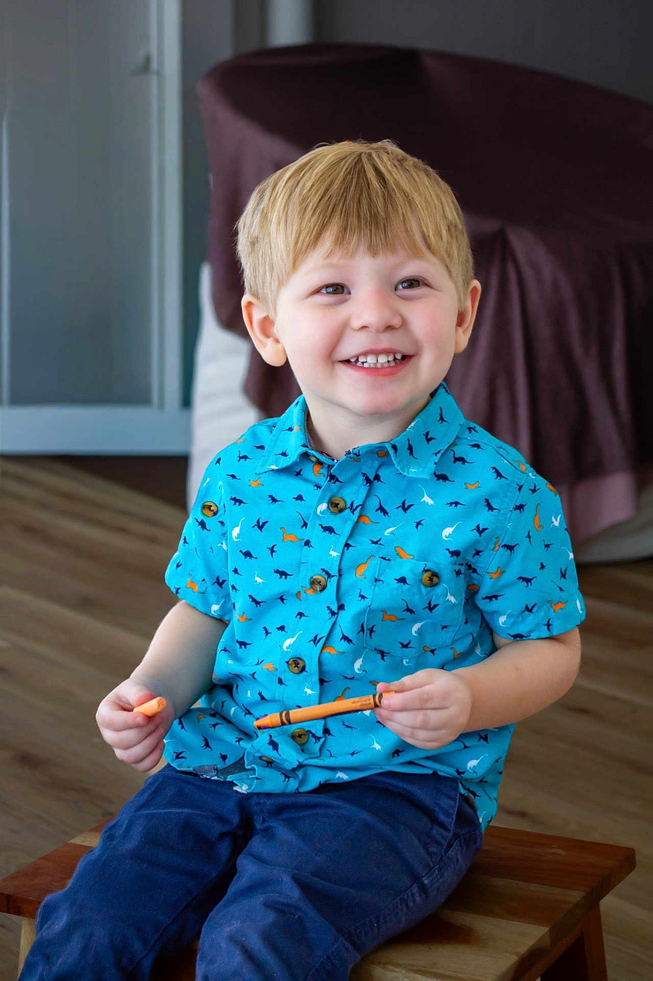 Ivan is registered to the contest to win money with this photo: child, boy, smiling, blue_shirt, dinosaur_pattern, crayon, sitting, wooden_bench, indoor, flooring, blonde_hair, happy, casual_clothing, portrait, playful, young, hands, teeth, face, eyes