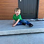 child, toddler, smiling, green_shirt, jeans, black_shoes, concrete, steps, door, wood_paneling, porch, happy, sitting, outside, young_child, face, person, casual, front_door, daylight