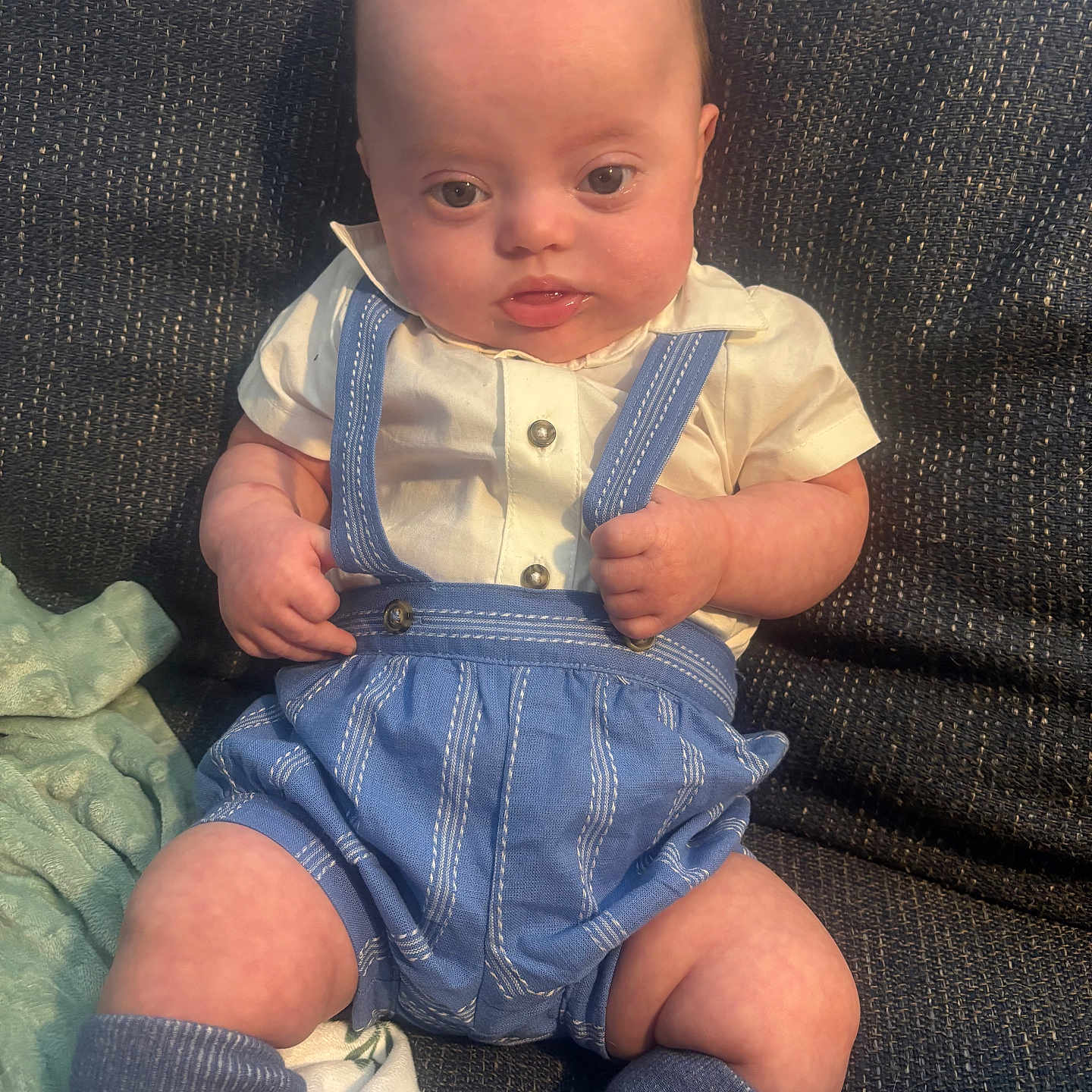 Warren is registered to the contest to win money with this photo: baby, infant, child, sitting, blue_overalls, cream_shirt, couch, textured_fabric, hands, legs, face, eyes, cute, indoors, natural_light, clothing, socks, expression, person, portrait