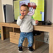 Ayden participe au concours pour gagner de l'argent avec cette photo : toddler, child, standing, casual_clothing, jeans, sneakers, television, cartoon, wooden_furniture, flooring, indoor, person, smiling, playful, modern, home, entertainment_unit, cable, white_sweatshirt, short_hair