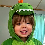 Elizabeth is registered to the contest to win money with this photo: child, toddler, portrait, smiling, happy, dinosaur_costume, green_hoodie, costume, hood, cartoon, teeth, face, eyes, hair, indoors, curtain, home, playful, cute, closeup
