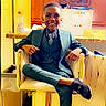 Camden is registered to the contest to win money with this photo: child, boy, smiling, suit, tie, chair, crossed_legs, indoor, kitchen, refrigerator, cabinet, countertop, plastic_bottle, happy, portrait, person, footwear, shoe, furniture, young_boy