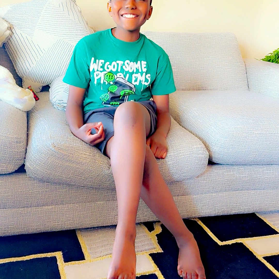 Camden joined the competition — help win amazing prizes! child, boy, smiling, sitting, couch, living_room, tshirt, barefoot, carpet, painting, home_interior, crossed_legs, happy, casual_clothing, shorts, furniture, indoor, person, portrait, relaxed