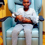 Camden is registered to the contest to win money with this photo: child, baby, armchair, blue_chair, indoor, flooring, wooden_floor, smile, boy, casual_clothing, sneakers, holding_baby, cozy, comfortable, person, seated, happy, family, home, portrait