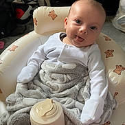 Mateo is registered to the contest to win money with this photo: baby, infant, blanket, chair, teddy_bear, container, tongue_out, white_clothing, indoor, sneakers, floor, carpet, furniture, cute, smiling, person, portrait, cozy, playful, baby_bottle_container