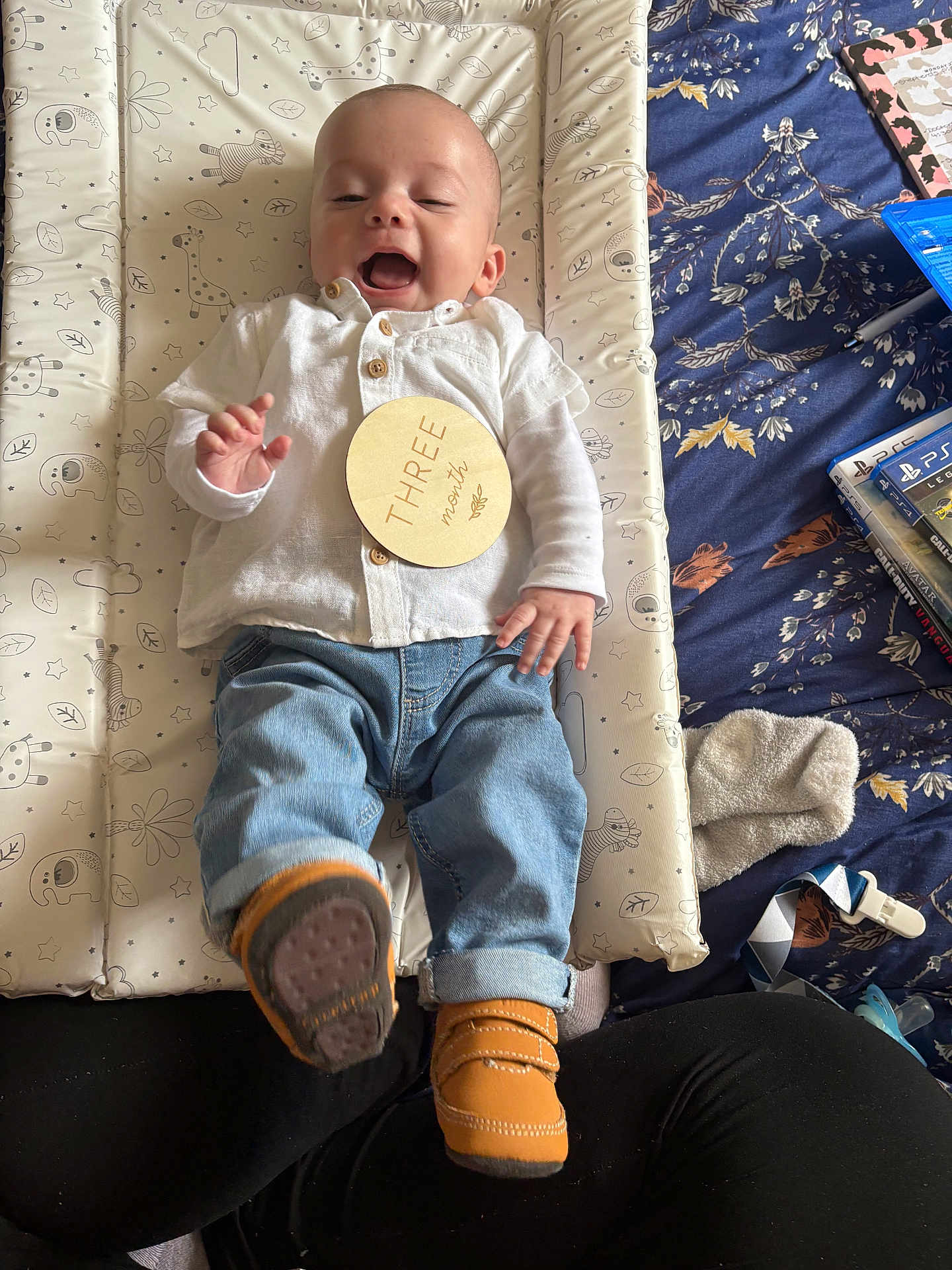 Mateo is registered to the contest to win money with this photo: baby, blue_blanket, changing_pad, clothing, cozy, face, hand, happy, infant, jeans, lying_down, milestone_card, pacifier, patterned, person, shirt, shoes, smiling, socks, video_games