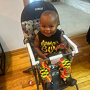 Boaz is registered to the contest to win money with this photo: baby, child, high_chair, smiling, indoor, wooden_floor, toy, clothing, colorful_pants, black_shirt, happy, person, footwear, plastic, home, furniture, seat, cute, expression, hand