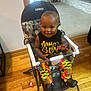 baby, child, high_chair, smiling, indoor, wooden_floor, toy, clothing, colorful_pants, black_shirt, happy, person, footwear, plastic, home, furniture, seat, cute, expression, hand