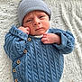 baby, beanie, blanket, blue_sweater, close_up, cozy, cute, expressive_face, hat, indoors, infant, knitwear, lying_down, newborn, portrait, sleepwear, small_hands, soft_texture, warm_clothing, wooden_buttons