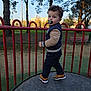 child, toddler, pacifier, playground, railing, park, trees, bench, apartment_building, urban, outdoor, curly_hair, jacket, sneakers, platform, daytime, grass, person, portrait, fence
