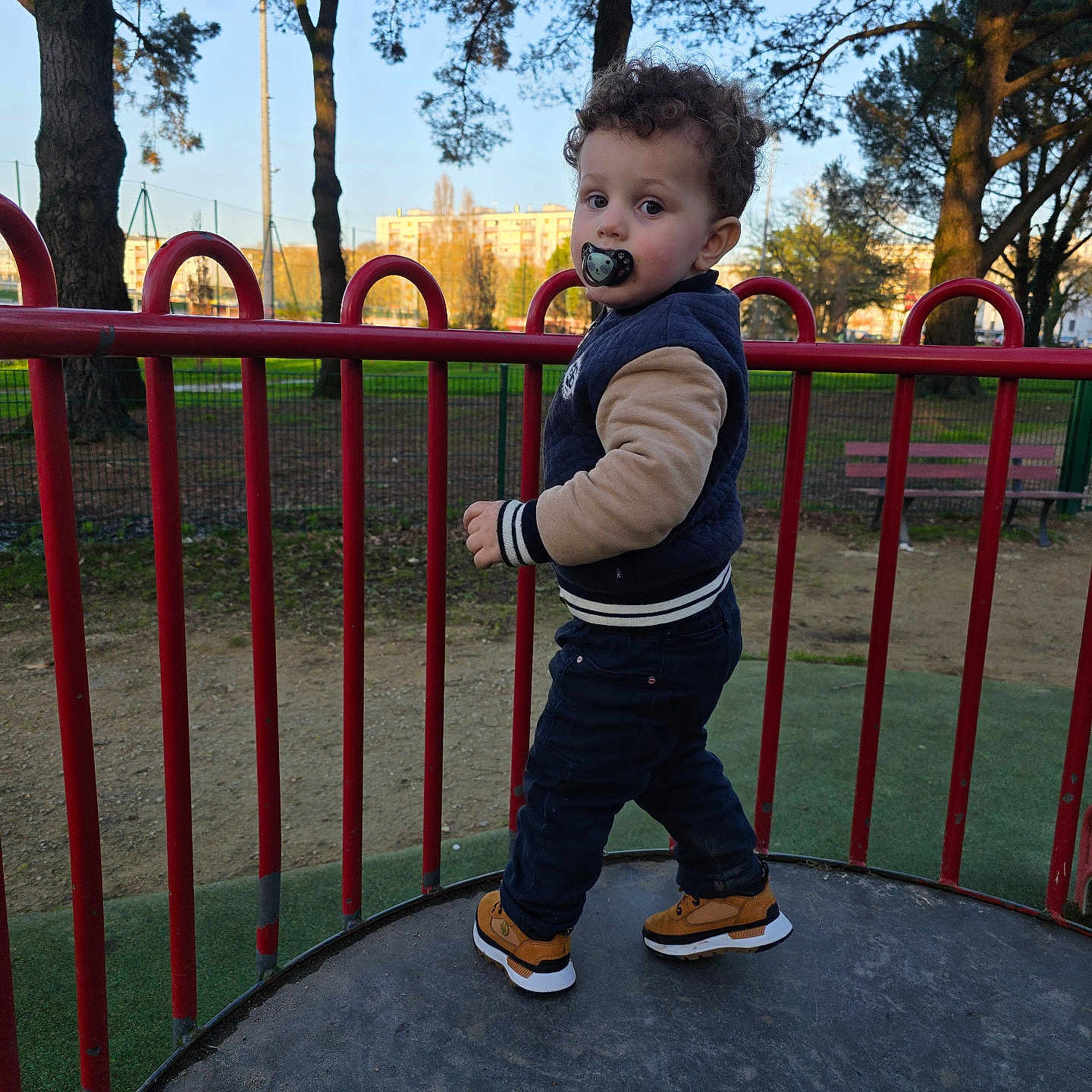 Aylan a rejoint le concours — aidez-le/la à gagner de superbes lots ! apartment_building, bench, child, curly_hair, daytime, fence, grass, jacket, outdoor, pacifier, park, person, platform, playground, portrait, railing, sneakers, toddler, trees, urban