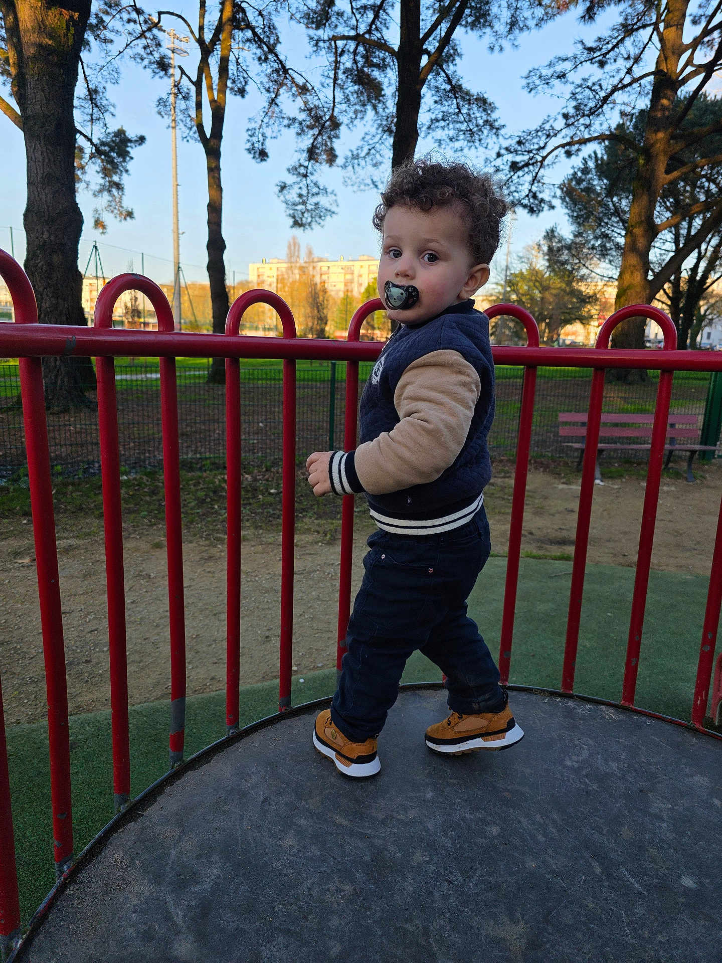 Aylan a rejoint le concours — aidez-le/la à gagner de superbes lots ! child, toddler, pacifier, playground, railing, park, trees, bench, apartment_building, urban, outdoor, curly_hair, jacket, sneakers, platform, daytime, grass, person, portrait, fence