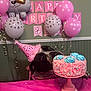 dog, birthday, party_hat, cake, pink, balloons, celebration, table, sprinkles, decorations, indoor, festive, animal, pet, party, pink_tablecloth, banner, paw_print, licking, cute