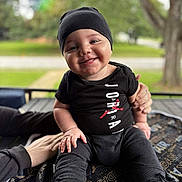 Jayden is registered to the contest to win money with this photo: baby, child, smile, black_clothing, hat, outdoor, greenery, blanket, sitting, happy, footwear, hand, person, portrait, cute, infant, fashion, daylight, closeup, support