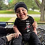 baby, child, smile, black_clothing, hat, outdoor, greenery, blanket, sitting, happy, footwear, hand, person, portrait, cute, infant, fashion, daylight, closeup, support