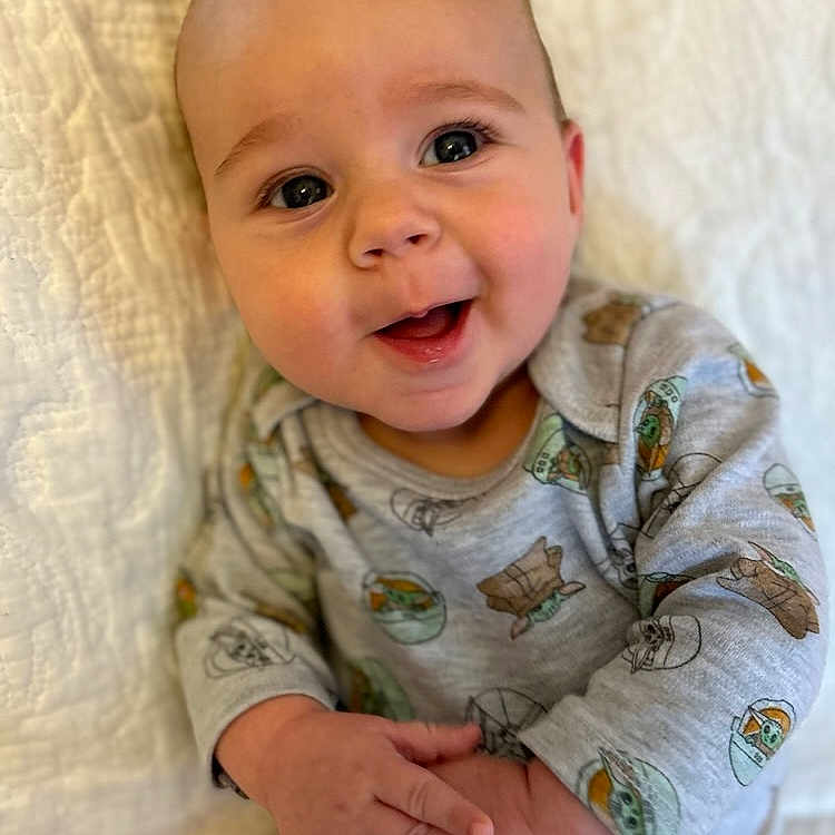 Hudson is registered to the contest to win money with this photo: baby, babylaughing, bodypart, clothing, face, finger, hand, happy, head, laughing, mouth, newborn, pajamas, person, photography, portrait, quilt, sitting, smile, teeth