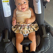 Sevyn joined the competition — help win amazing prizes! baby, child, car_seat, headband, skirt, beige_top, fuzzy_slippers, smiling, indoor, footwear, plaid_pattern, person, infant, cute, happy, seatbelt, floor, sneaker, fashion, portrait