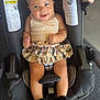 baby, child, car_seat, headband, skirt, beige_top, fuzzy_slippers, smiling, indoor, footwear, plaid_pattern, person, infant, cute, happy, seatbelt, floor, sneaker, fashion, portrait