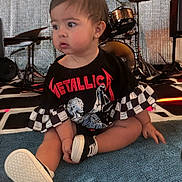 Sofia is registered to the contest to win money with this photo: baby, black_clothing, carpet, checkered_pattern, child, curious, cute, drum_set, earrings, floor, indoor, metallica, musical_instruments, person, rock_music, short_hair, sitting, sneakers, toddler, tshirt