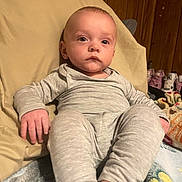 Elijah joined the competition — help win amazing prizes! baby, child, pajamas, gray_clothing, cushion, chair, indoor, wood_paneling, face, person, footwear, blanket, relaxing, infant, portrait, seated, hand, expressionless, home, cozy