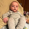 baby, child, pajamas, gray_clothing, cushion, chair, indoor, wood_paneling, face, person, footwear, blanket, relaxing, infant, portrait, seated, hand, expressionless, home, cozy