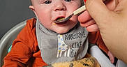Noah is registered to the contest to win money with this photo: activity_panel, adult_hand, baby, bib, blanket, child, close_up, eyes, face, feeding, high_chair, indoor, infant, meal, orange_clothing, plush_blanket, portrait, sitting, spoon, toy