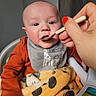 baby, infant, child, feeding, spoon, adult_hand, high_chair, bib, orange_clothing, blanket, plush_blanket, toy, activity_panel, indoor, portrait, close_up, face, eyes, sitting, meal