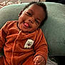 baby, child, smiling, tongue_out, orange_outfit, hedgehog_patch, cozy, cushion, indoor, cute, happy, person, portrait, soft_texture, infant, playful, furniture, relaxed, young_child, casual
