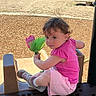 bench, casual_wear, child, curly_hair, cute, daylight, flower_toy, leisure, nature, outdoor, park, pink_clothing, play_equipment, playground, sand, shoes, sitting, sunlight, toddler, young_child