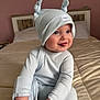baby, bed, bedding, blue_eyes, cheeky, child, cozy, cute, hand, hat, indoor, infant, knotted_hat, person, playful, portrait, ribbed_clothing, smiling, soft_lighting, tongue_out