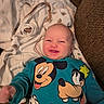 baby, smiling, mickey_mouse, donald_duck, goofy, sweater, blanket, couch, happy, child, infant, clothing, portrait, lying_down, cute, young_child, indoors, person, face, cozy